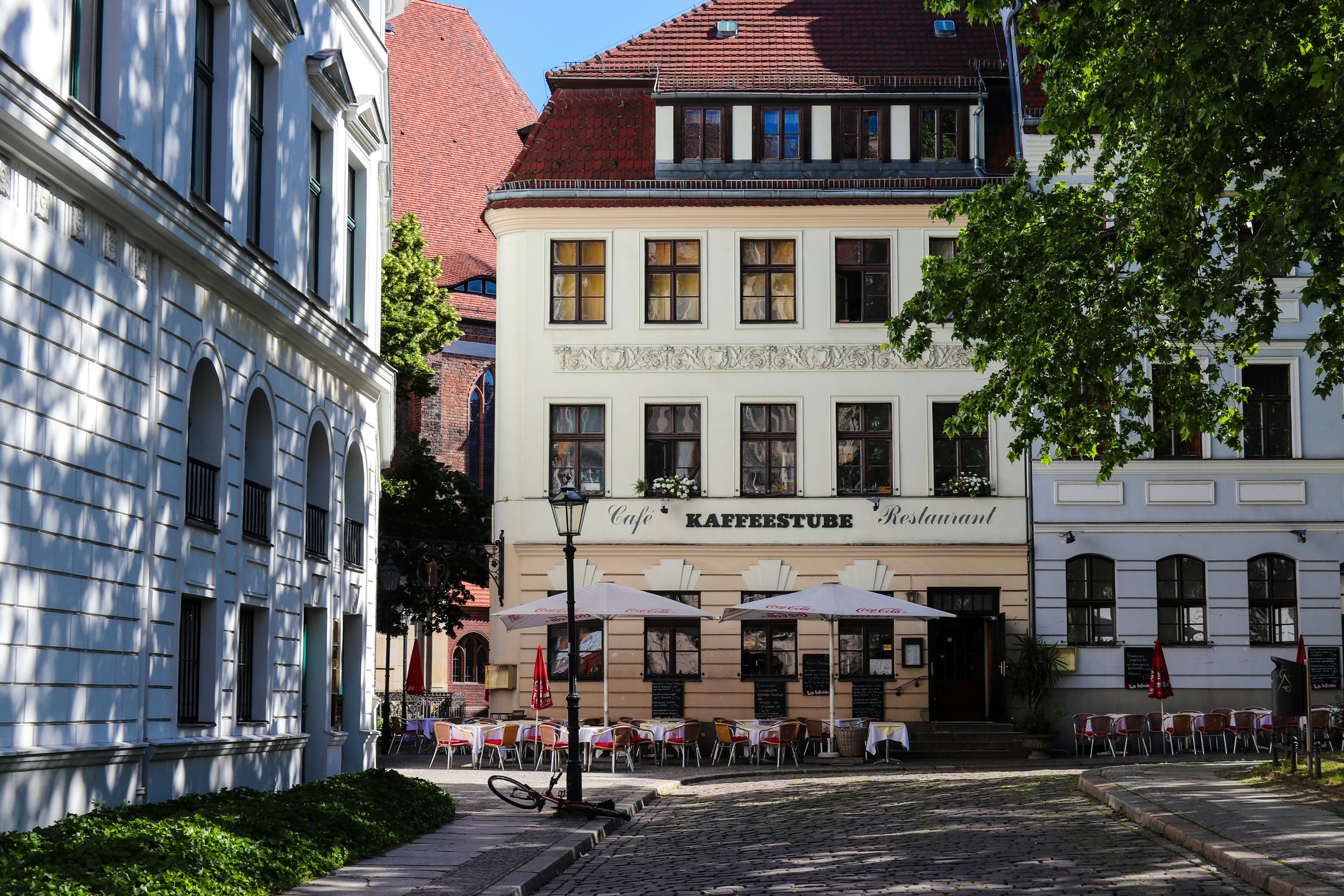 Außenansicht eines Restaurants mit Tischen und Stühlen im Freien, umgeben von historischen Gebäuden, eine große grüne Baum und eine schmale gepflasterte Straße. Auf dem Gebäude ist ein Schild mit der Aufschrift "Kaffeestube".