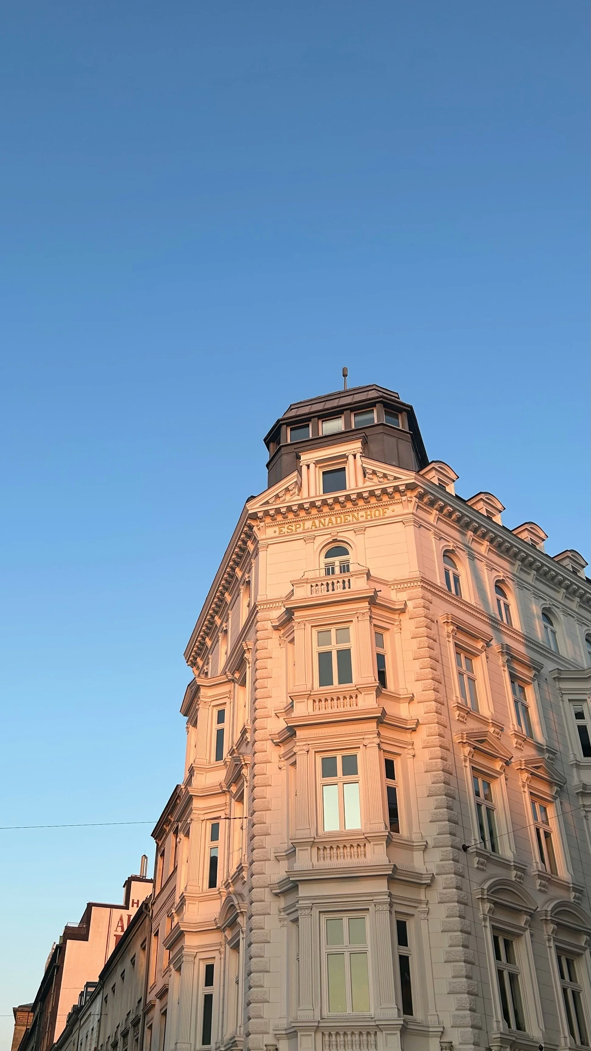 Ein mehrstöckiges historisches Gebäude mit barocker Architektur, das den Namen 'ESPLANADE-HOF' trägt, steht gegen einen blauen Himmel, während die Abendsonne das Gebäude in warmes Licht taucht.