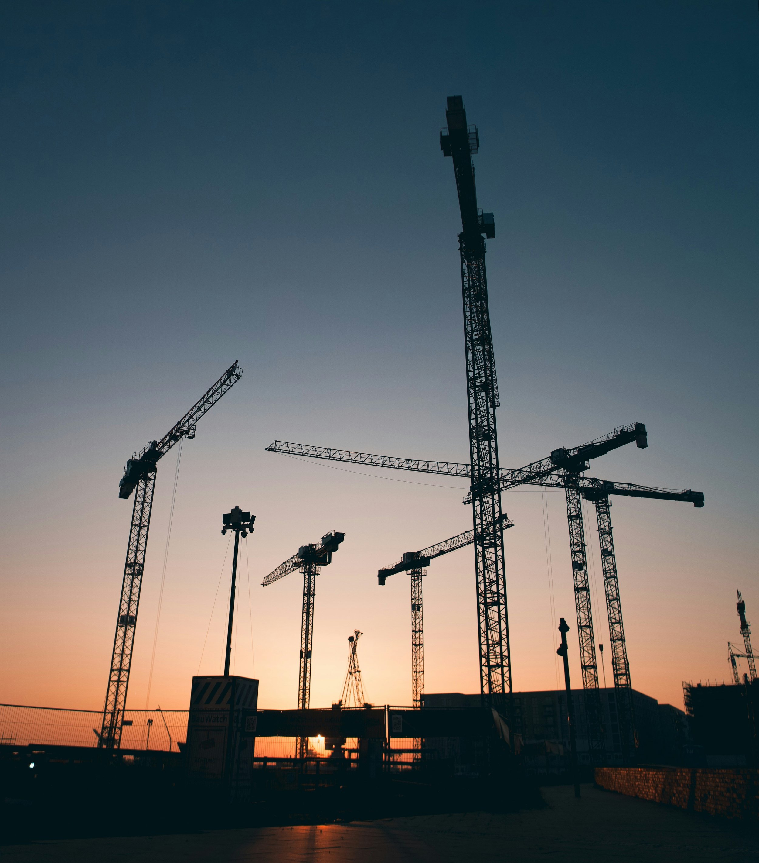 Baukräne auf einer Baustelle während des Sonnenuntergangs, mit dunklem Himmel im Hintergrund.