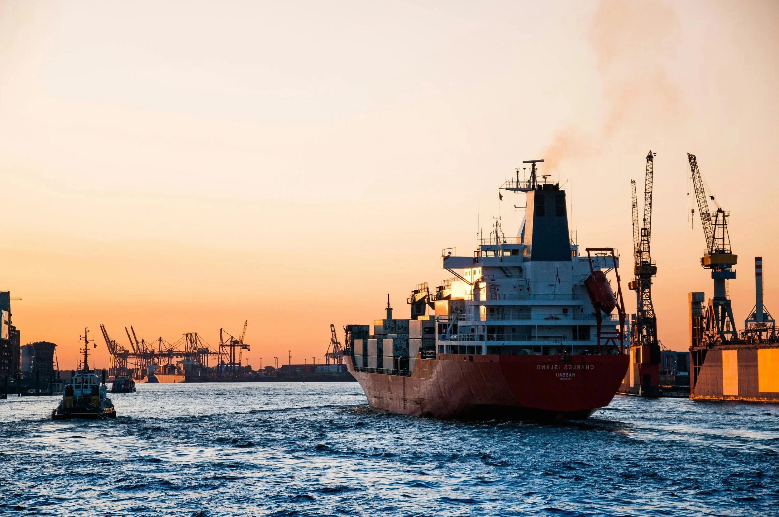 Ein Frachtschiff namens Chelsea Sand in einem Hafen bei Sonnenuntergang, umgeben von Kränen und kleineren Booten auf dem Wasser.
