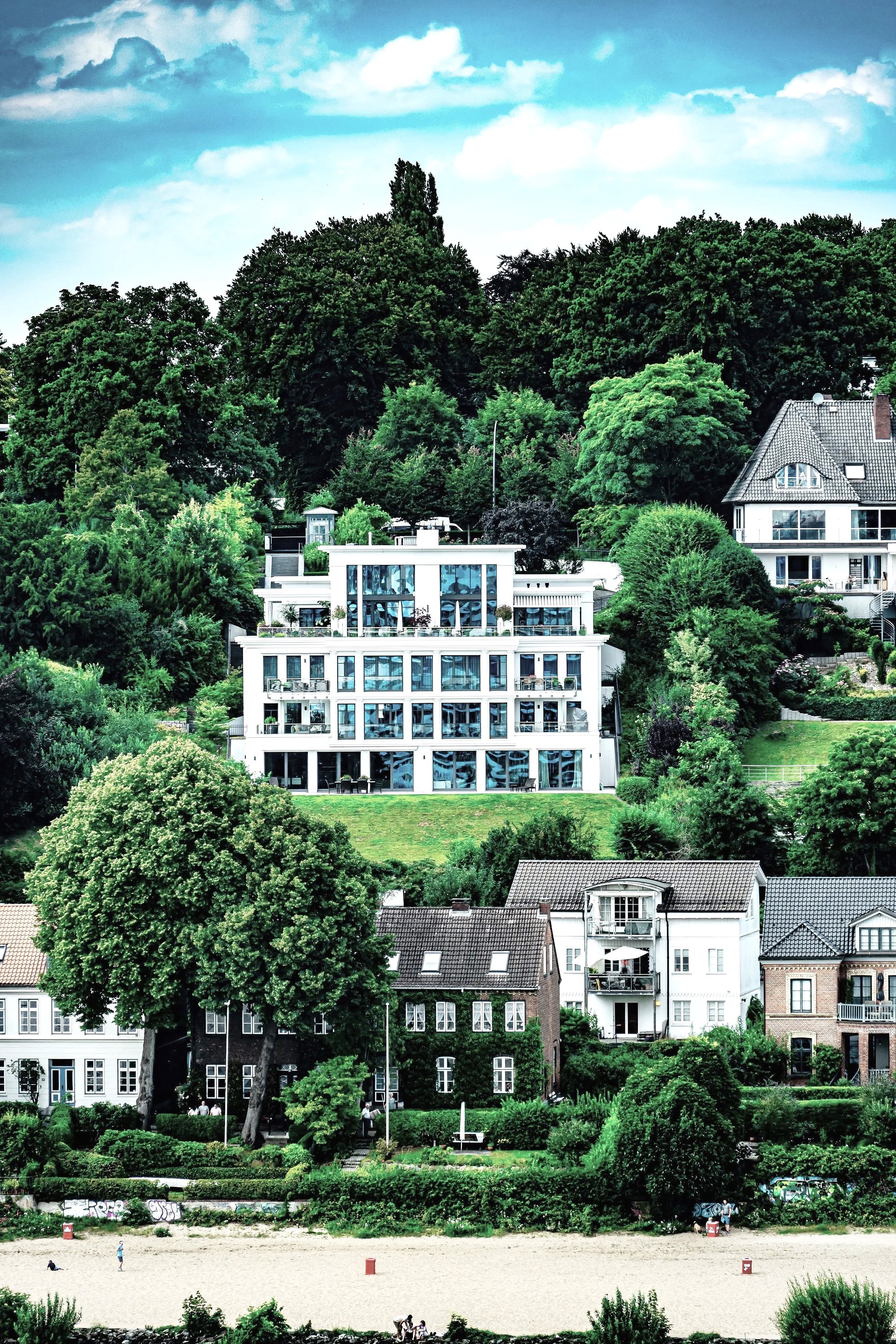 Mehrstöckiges modernes weißes Gebäude mit großen Fenstern, umgeben von vielen Bäumen und grünem Garten, an einem Hang in einer städtischen Nachbarschaft, im Hintergrund ältere Häuser mit Ziegeldächern, vor einem bewölkten Himmel.