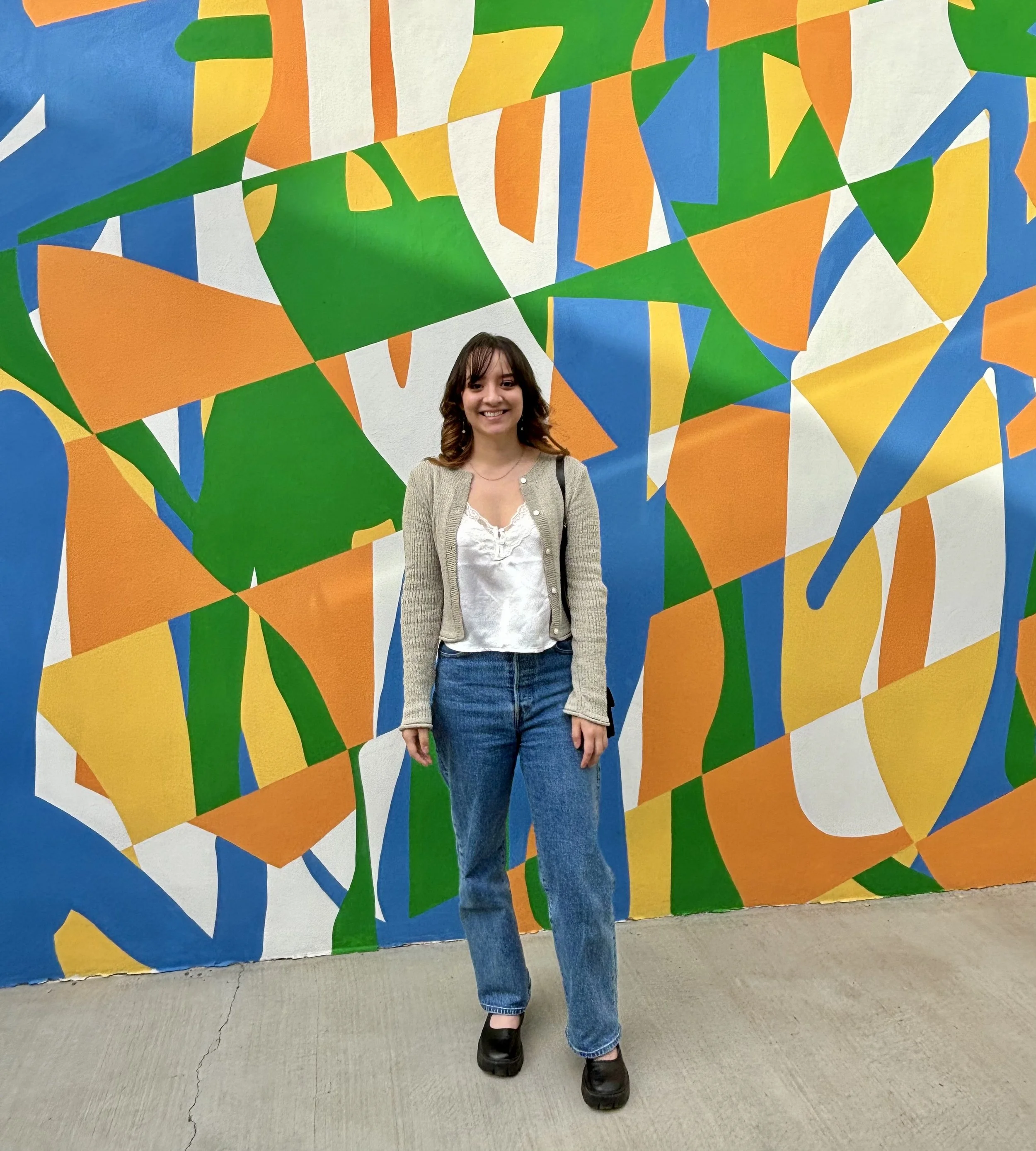 A young woman with shoulder-length brown hair, wearing a beige cardigan, white blouse, and blue jeans, stands in front of a colorful abstract mural with green, orange, yellow, white, and blue geometric shapes.