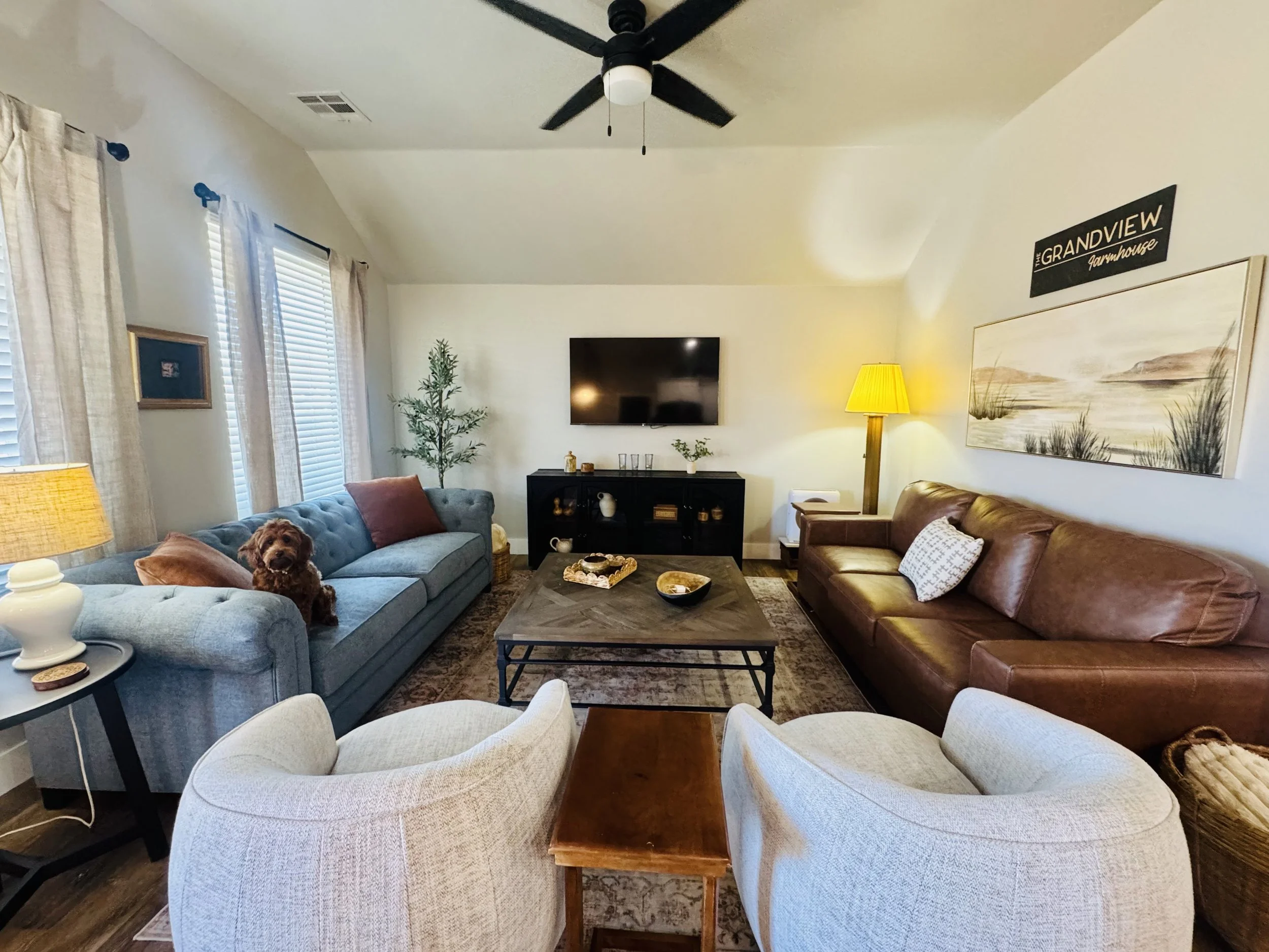 Spacious living room at Grandview Farmhouse in Stillwater, Oklahoma featuring blue sectional sofa, brown leather couch, cozy seating, wall-mounted TV, warm lighting, and pet-friendly vibe with a friendly dog
