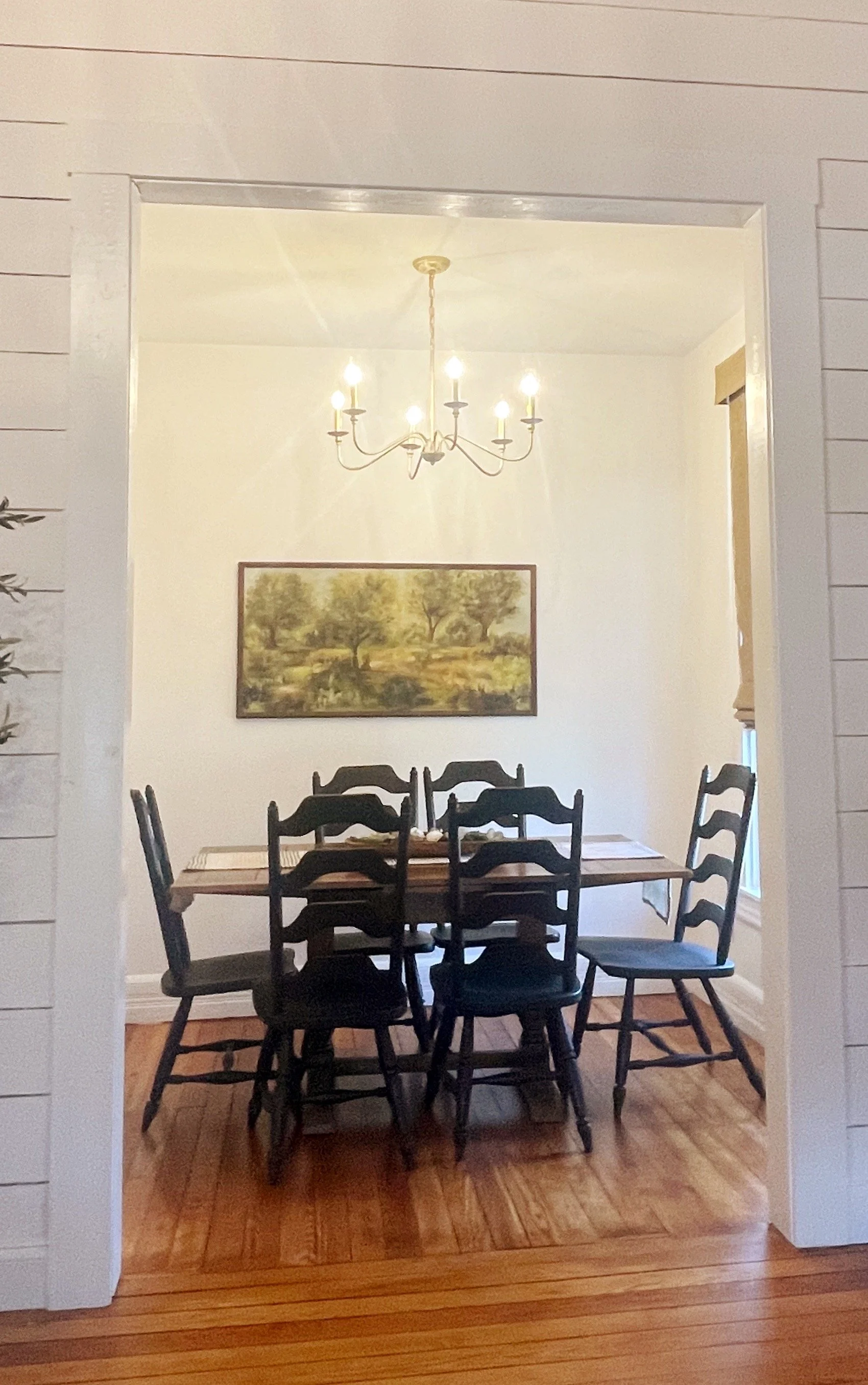 Charming dining room at Clay Oak Cottage in Waco, Texas featuring rustic wooden table, black ladder-back chairs, gold chandelier, and large landscape painting of trees