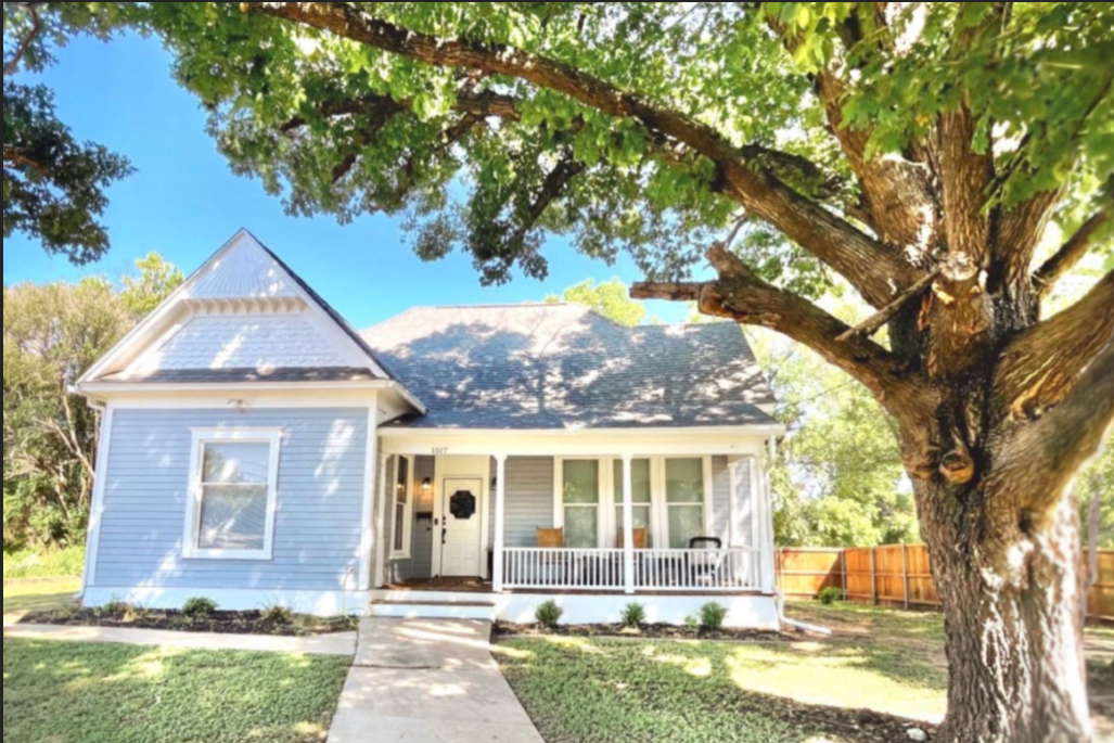 Charming exterior of Clay Oak Cottage in Waco, Texas – light blue farmhouse with classic front porch, white columns, rocking chairs, and shady oak trees