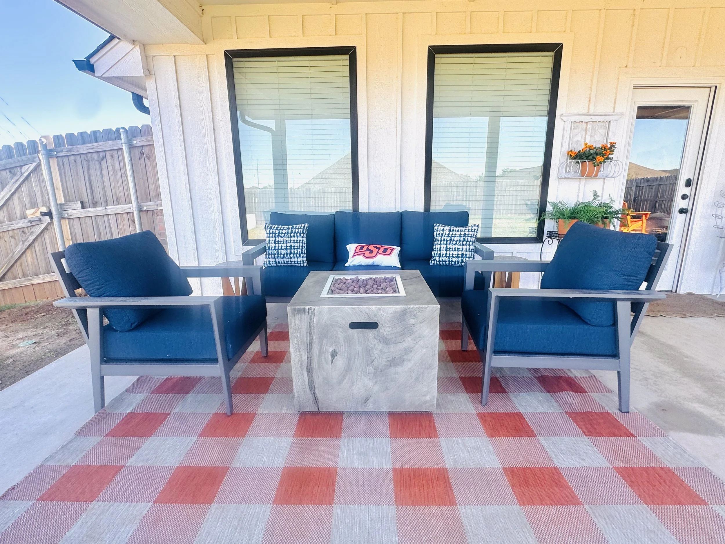 Cozy covered back porch at Grandview Farmhouse in Stillwater, Oklahoma with navy blue outdoor sofa, armchairs, gas fire pit table, and OSU pillow