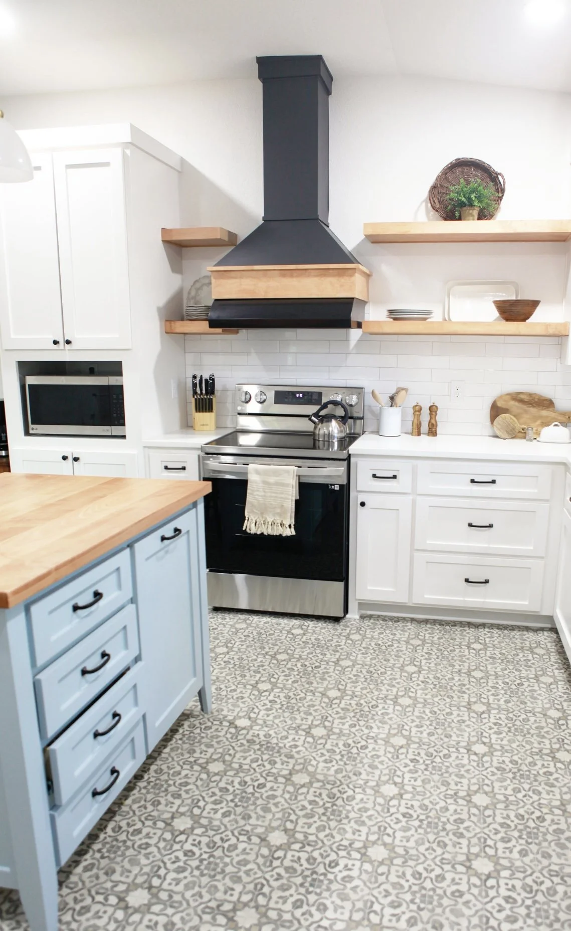 Bright modern farmhouse kitchen at Clay Oak Cottage in Waco, Texas featuring white cabinets, blue island, butcher block counters, black range hood, subway tile backsplash, and open shelving