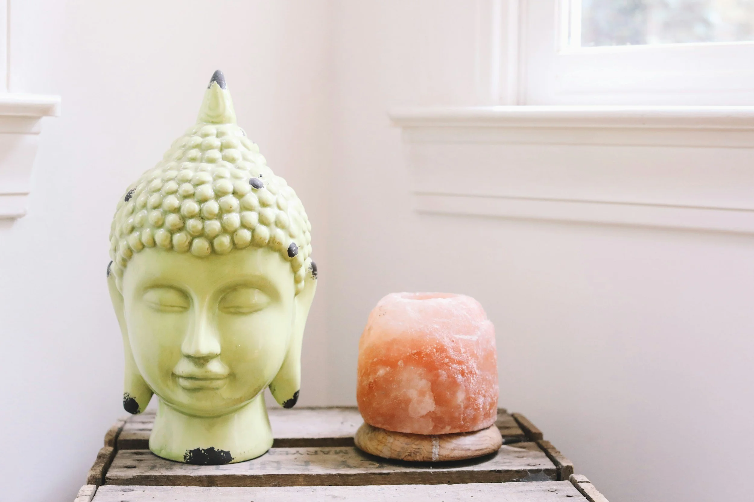 A ceramic Buddha head sculpture beside a pink Himalayan salt lamp on a wooden surface indoors with a window in the background.