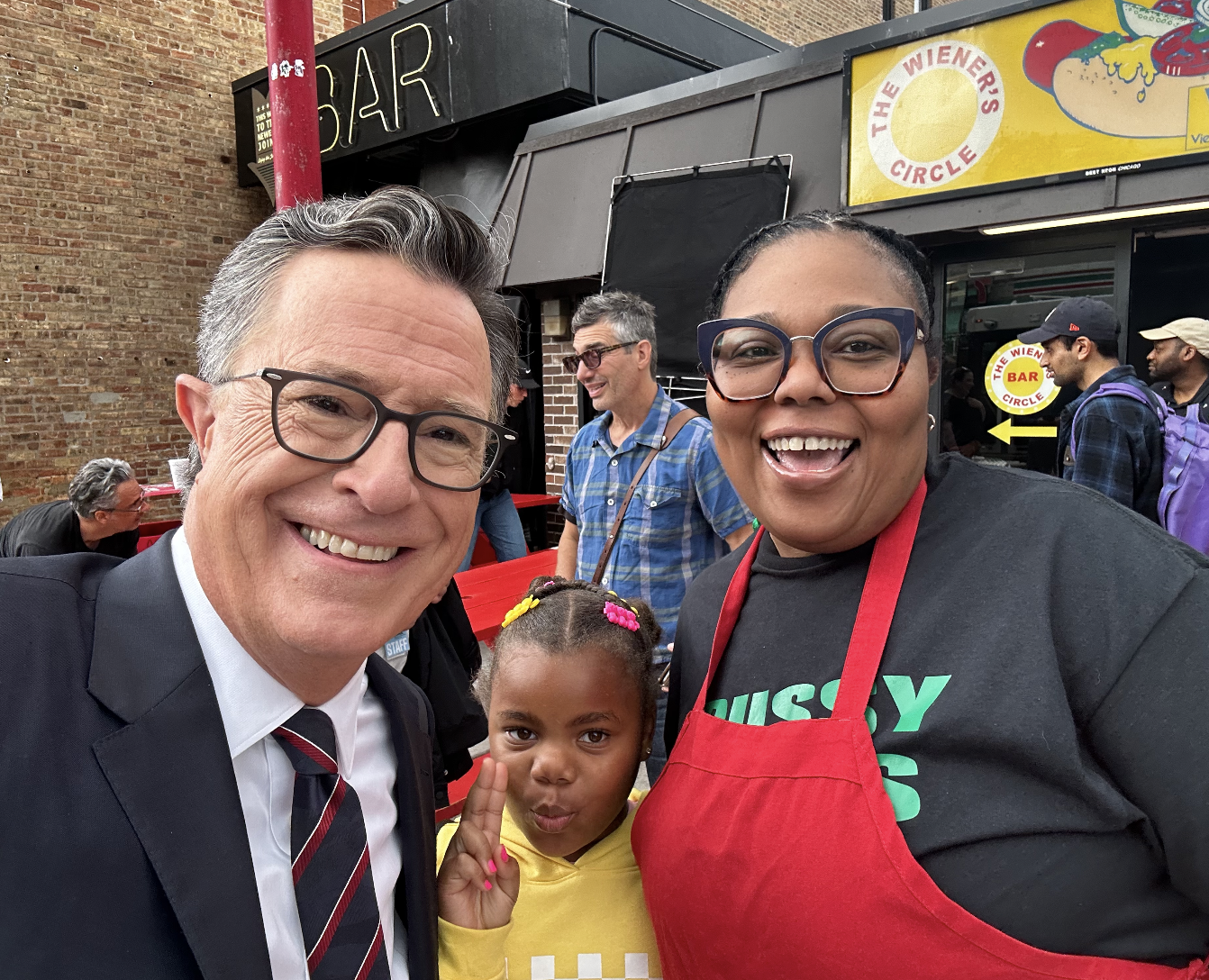 Three smiling people, two adults and one child, taking a selfie in front of a food stand. The man on the left is wearing glasses and a business suit with a tie. The woman on the right is wearing glasses, a black T-shirt, and a red apron. The girl in 