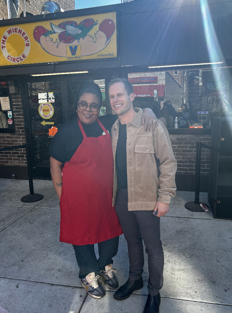 Two people standing together outside a restaurant called The Wiener's Circle. The woman on the left is wearing glasses, a red apron, and sneakers, with her arm around the man. The man on the right is smiling, wearing a beige jacket, black pants, and 