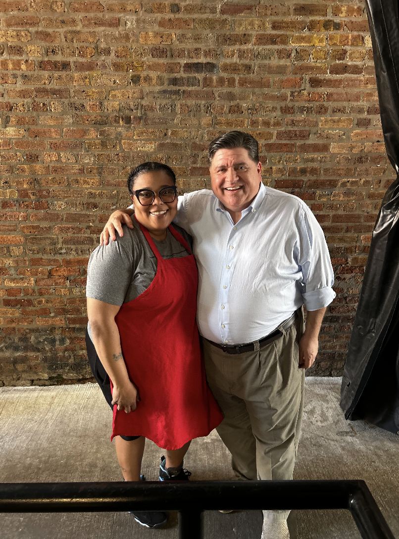 A woman wearing glasses and a red apron stands next to a man in a white shirt. They are smiling and standing close together against a brick wall.