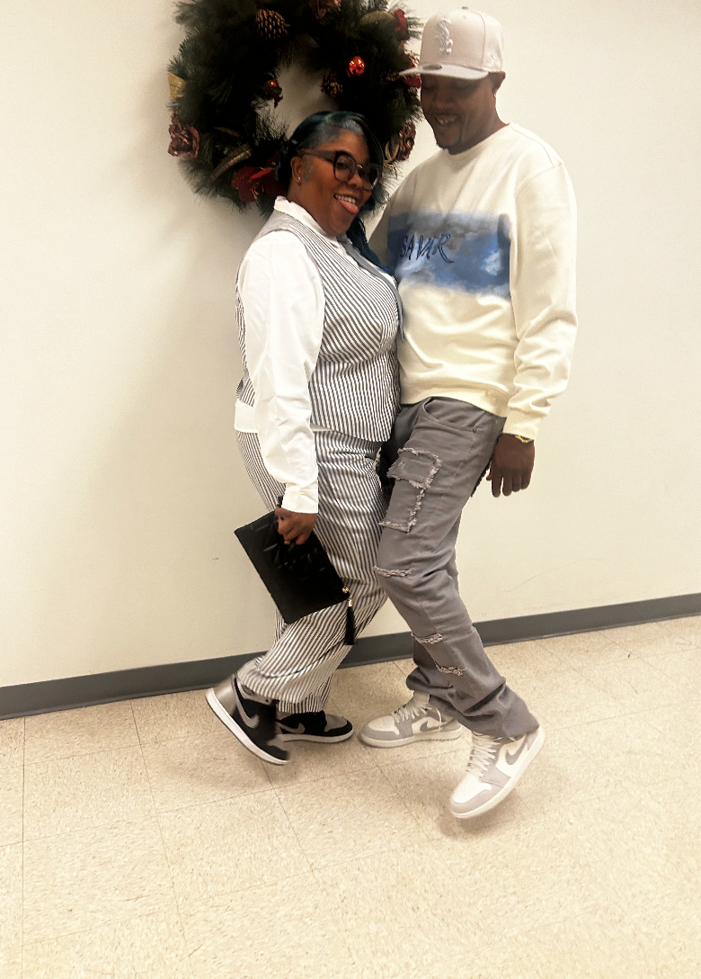 A woman and a man are posing together indoors by a wall decorated with a Christmas wreath. The woman is wearing black glasses and a striped outfit, holding a black clutch purse. The man is wearing a white cap, a white sweatshirt, and gray distressed 