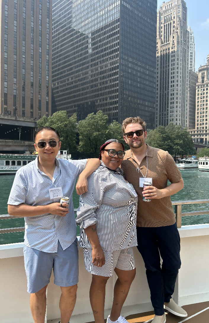 Three friends posing together outdoors on a boat cruise in a city with tall skyscrapers, holding drinks and smiling at the camera.