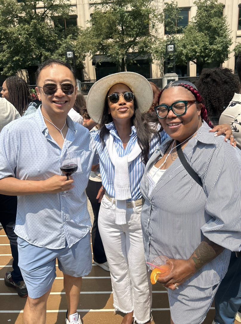 Three friends enjoying an outdoor gathering on a sunny day, with trees and a building in the background. They are smiling and holding drinks, dressed casually with sunglasses and hats.