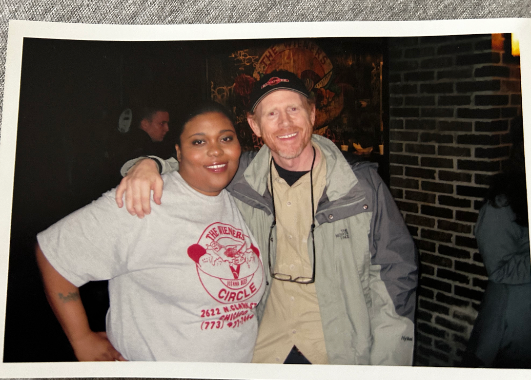 A woman with dark hair and makeup, and a man with light hair and a hat, posing together in a dimly lit bar or club. They are smiling, with the man's arm around the woman's shoulders.