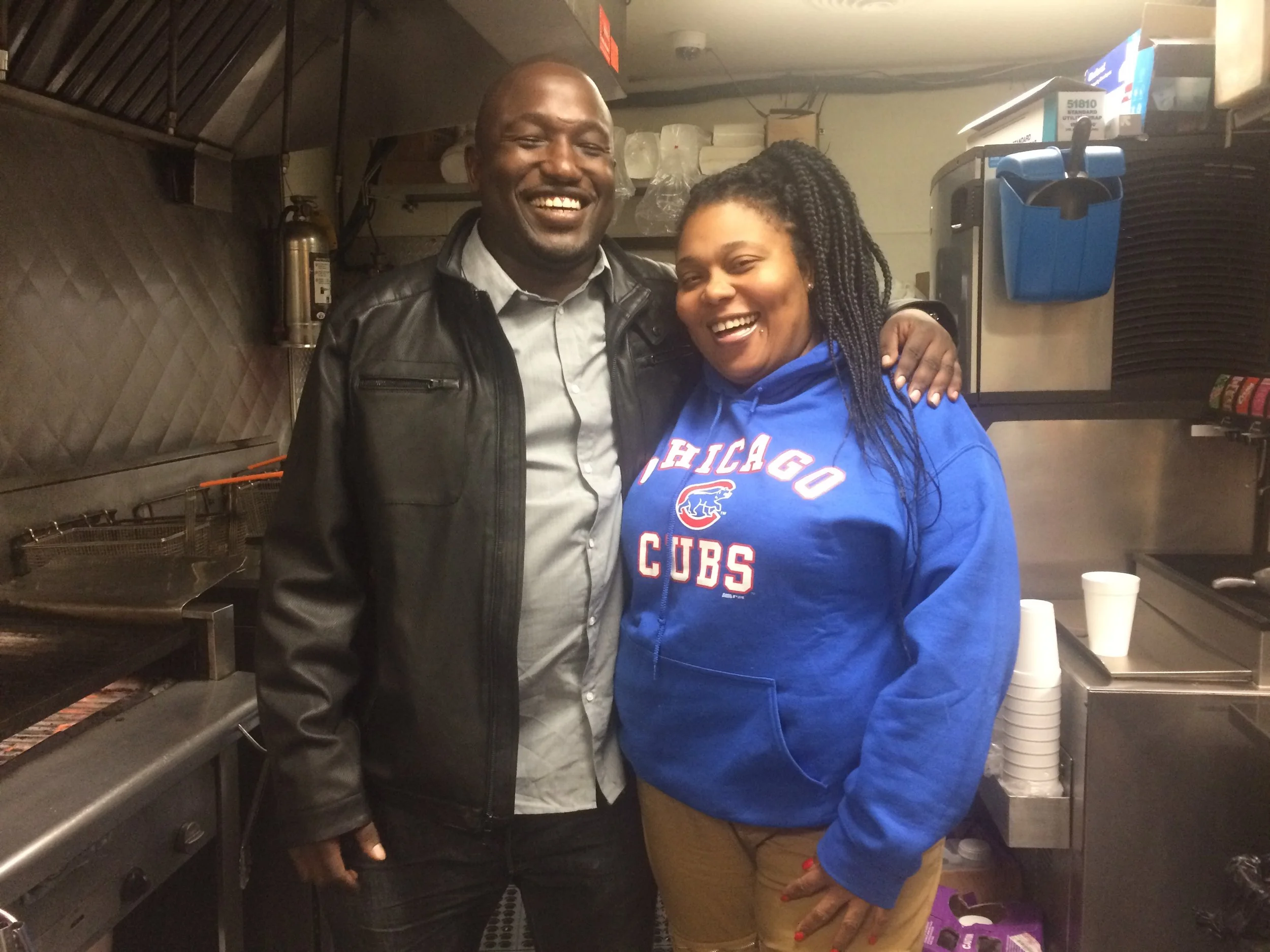 A man and woman smiling and standing together in a restaurant kitchen. The man wears a black leather jacket and the woman wears a blue Chicago Cubs hoodie.