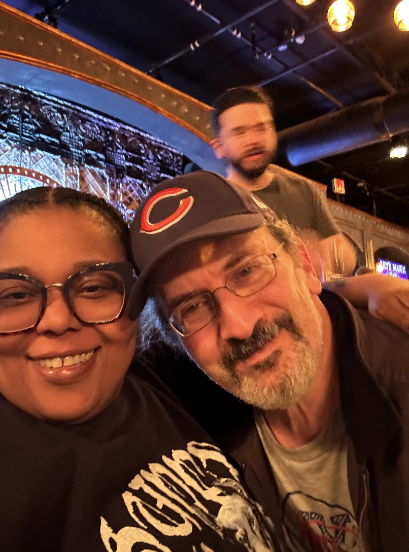 A smiling woman with glasses and a man with glasses and a baseball cap taking a selfie in a lively indoor setting, with another man in the background.
