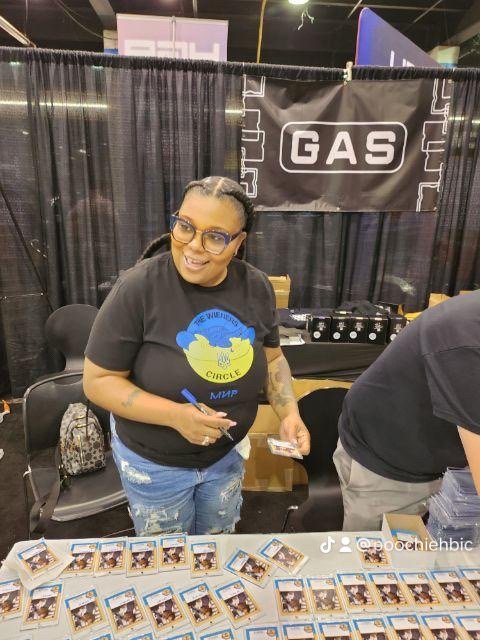 Woman with glasses, black t-shirt, and tattoos, standing behind a table with photo cards at an event booth.
