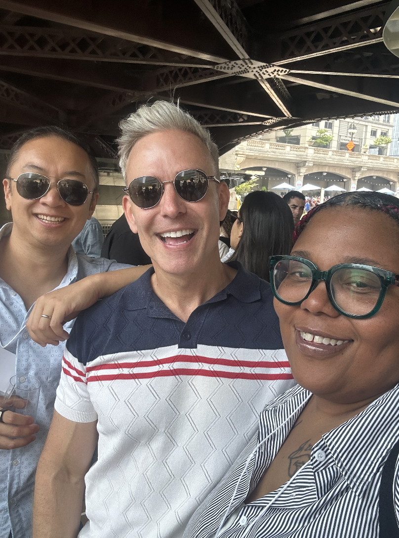 Three people taking a selfie under a bridge, smiling, with other people in the background.