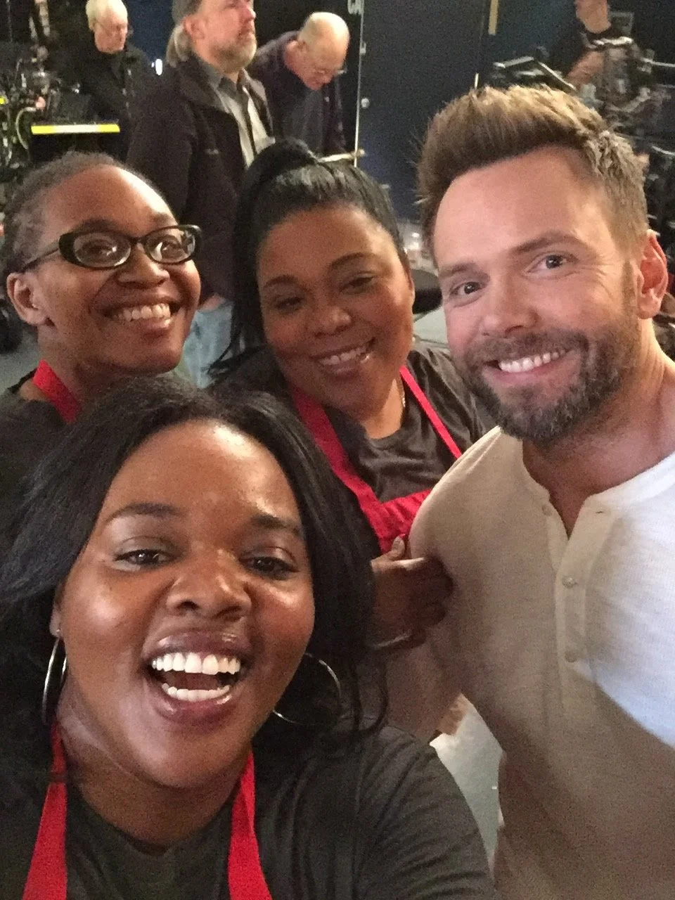 Group of four smiling people taking a selfie in a busy indoor setting, with other individuals and equipment in the background.
