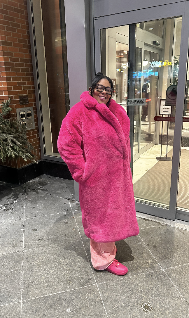 A smiling woman in a bright pink, fuzzy coat, pink pants, and pink shoes standing outside in front of a glass entrance door to a building at night.