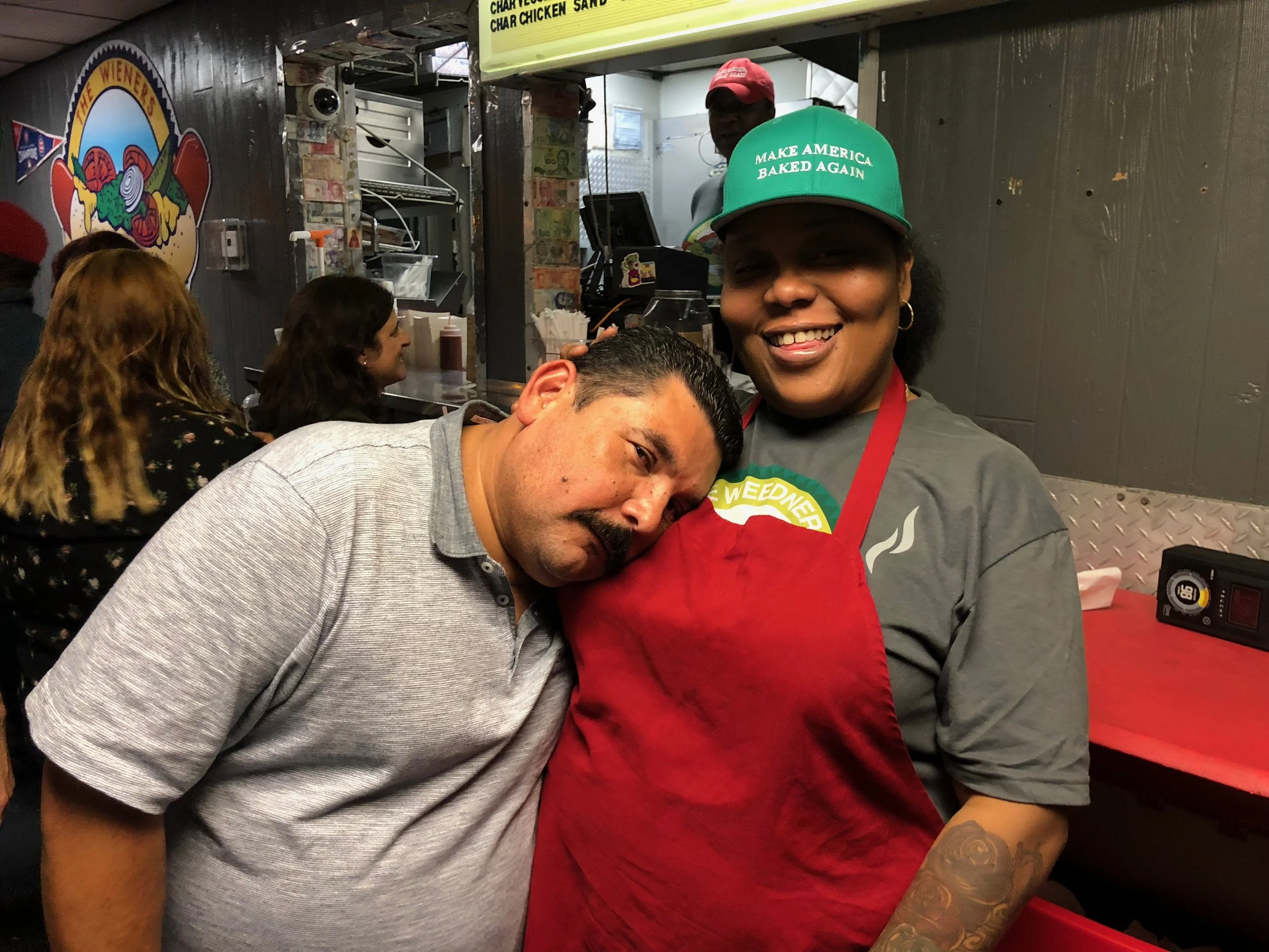 A woman wearing a green cap that says 'Make America Baked Again,' smiling, standing behind a man with a mustache who is resting his head on her chest. They are inside a casual restaurant, with other people in the background and a sign above them.