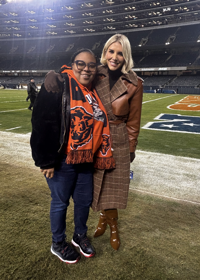 Two women standing on a football field at night, smiling with their arms around each other, wearing warm clothing. One is dressed in a brown plaid coat and high boots, and the other in a black jacket, orange scarf and glasses.