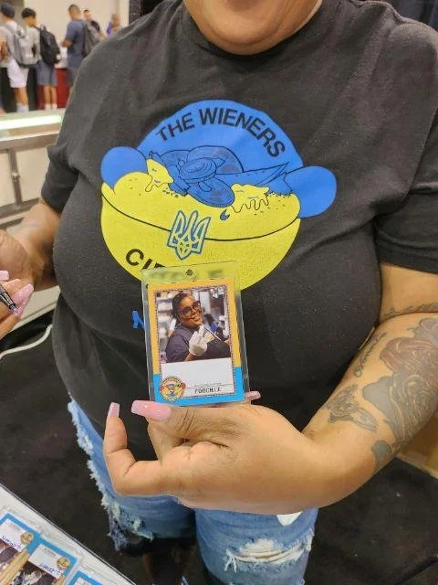 Person holding a Poodie trading card at a convention, wearing a gray T-shirt with a cartoon logo of a hot dog and a blue bird, with a yellow Ukrainian trident symbol on the shirt.