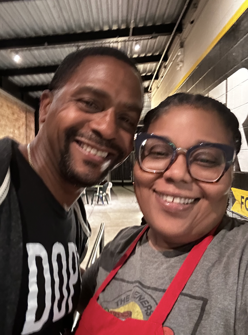 A smiling man and woman taking a close-up selfie in a warehouse or industrial kitchen setting. The woman is wearing glasses and a red apron, and the man is wearing a black shirt with white text.