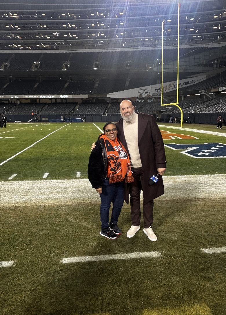 Two people smiling and posing on a football field at a stadium, with goalposts and empty stands in the background.