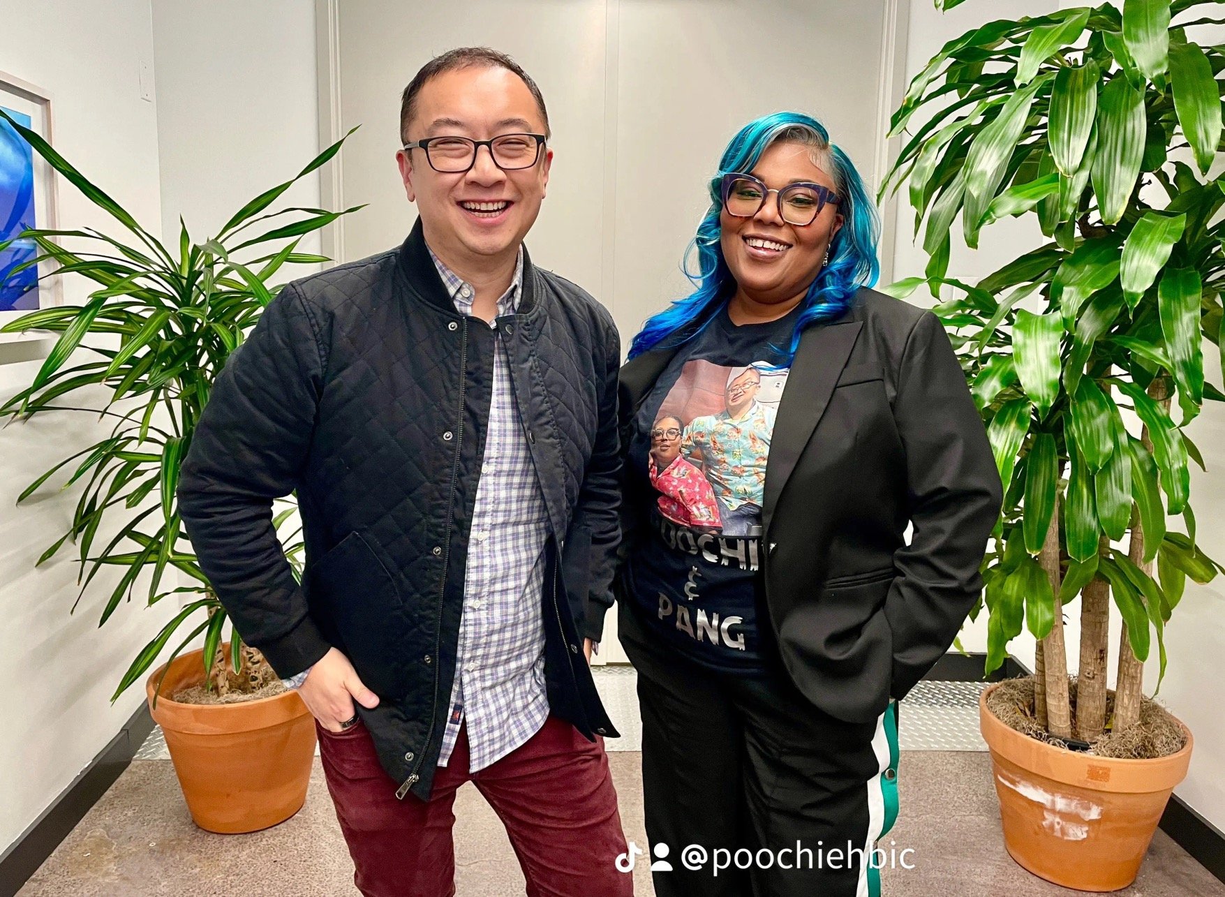 A smiling man with glasses wearing a black quilted jacket and plaid shirt, standing next to a smiling woman with blue hair and glasses, wearing a black blazer and a graphic T-shirt, in an office with large potted plants.
