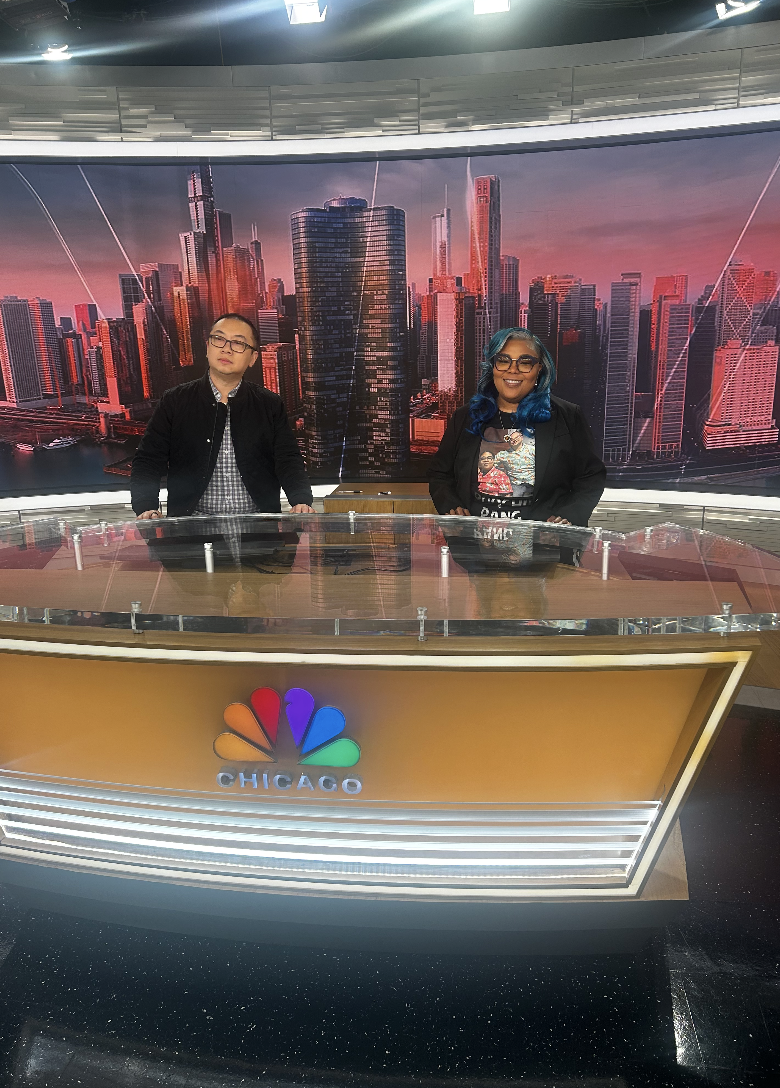 Two news anchors, a man and a woman, standing behind a news desk with the NBC Chicago logo, with a cityscape of Chicago skyline in the background.