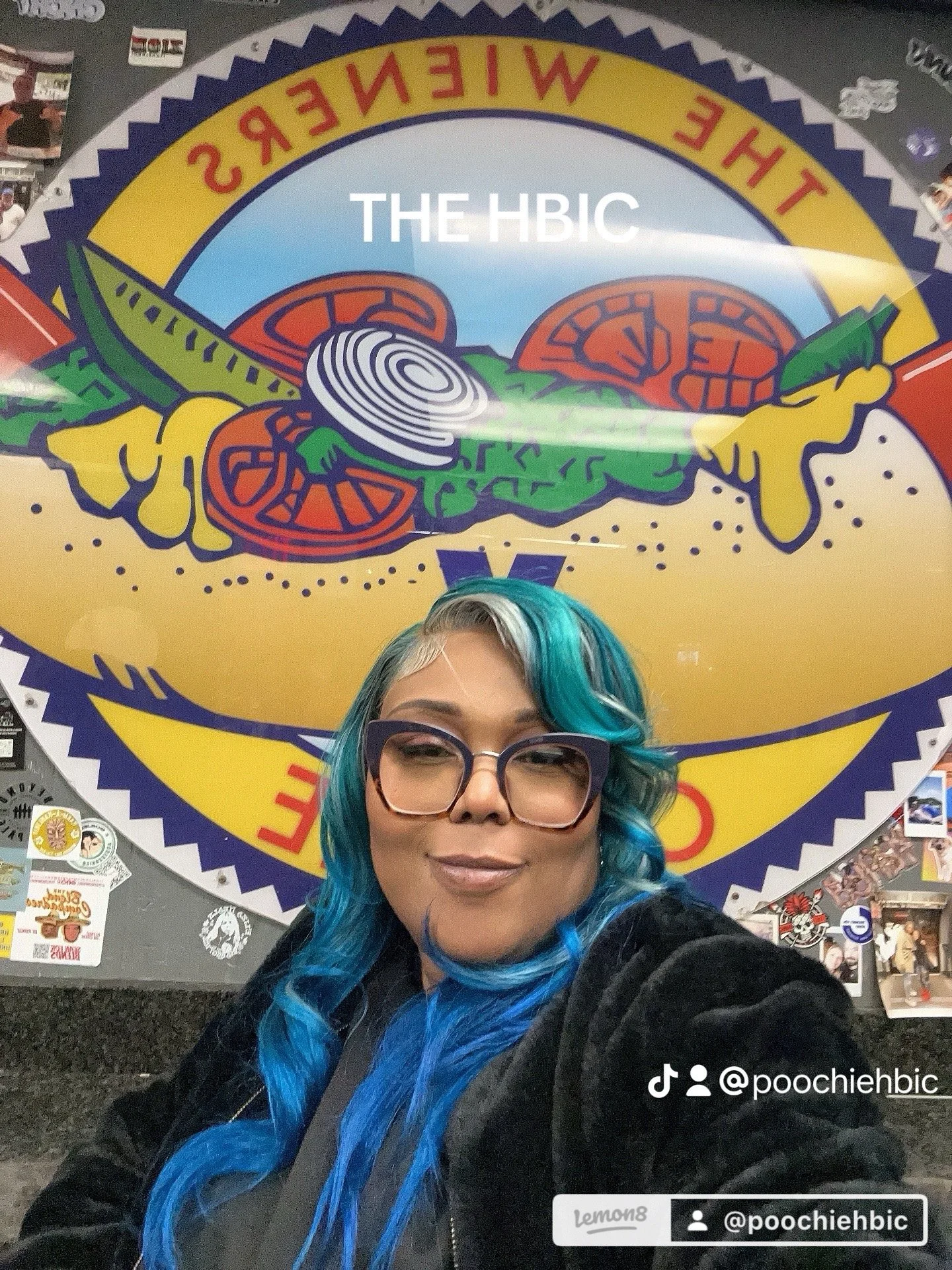 A woman with colorful blue hair and glasses taking a selfie in front of a large circular sign that reads 'The HBIC' with a graphic of a joint and vegetables.