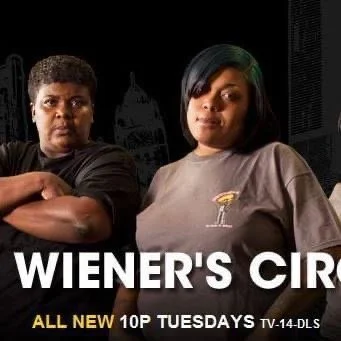 Two women sitting in a dark room with a cityscape in the background, one with crossed arms and the other with hands on hips, promoting Wiener’s Circle TV show.