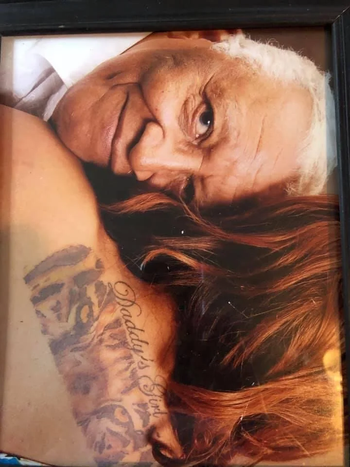 A close-up photo of an elderly woman with gray hair lying with her head resting on a woman's shoulder, whose long red hair partially covers her face and a visible tattoo on her shoulder.