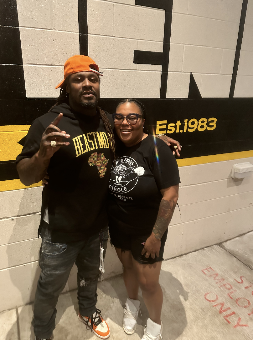 Two people standing together in front of a painted wall with the text 'Est. 1983'. The person on the left is a man wearing an orange baseball cap backward, a black hoodie with 'BEASTING' and an African continent graphic, ripped jeans, and sneakers, m