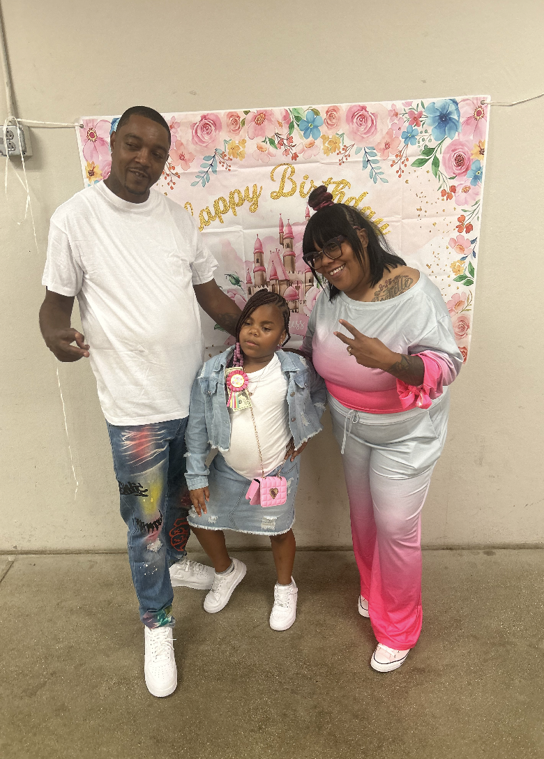 Family of three celebrating birthday in front of a floral birthday banner with a castle, with the child in the center and adults on either side.