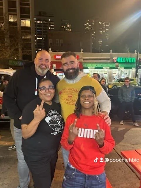 Four people smiling and posing on a city street at night, with a 7-Eleven store in the background. Two women and two men, some making hand gestures.