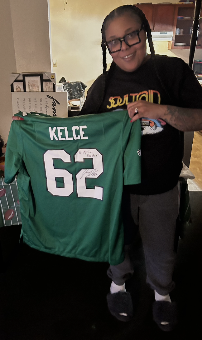 Woman smiling, holding green sports jersey with the name Kelce and the number 62, signed and personalized with a note that says 'To My Girl Poochi!!' in a cozy room.