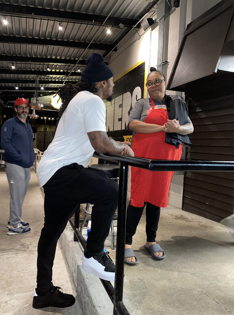 A man with long dreadlocks and a beard, wearing a black beanie, white t-shirt, black pants, and white sneakers, talking to a woman in a red apron and glasses, inside a restaurant or food establishment.