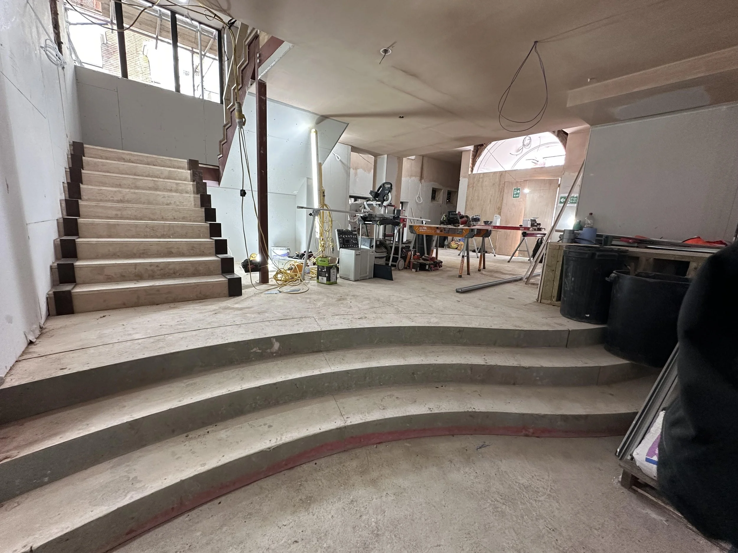 Interior of a building under construction with curved steps, a staircase leading up, construction tools, and materials scattered around, with unfinished walls and an opening allowing natural light.