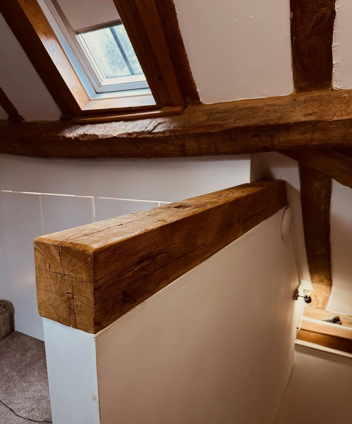 Close-up of a wooden handrail on a staircase, with a wood-beamed ceiling and a window letting in natural light.