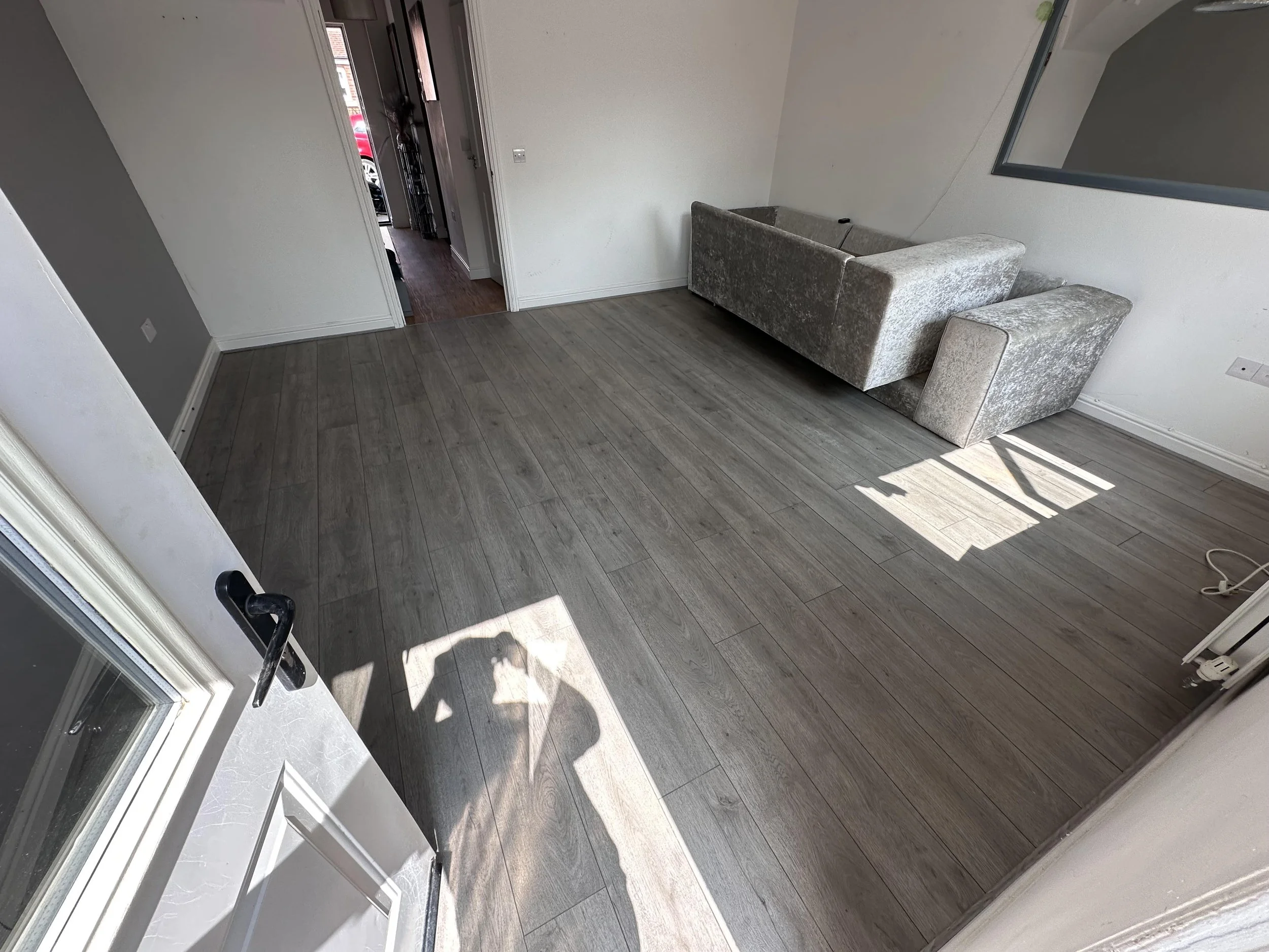 Empty living room with grey wooden floor and a grey sofa near the wall. Sunlight casting shadows through a window.