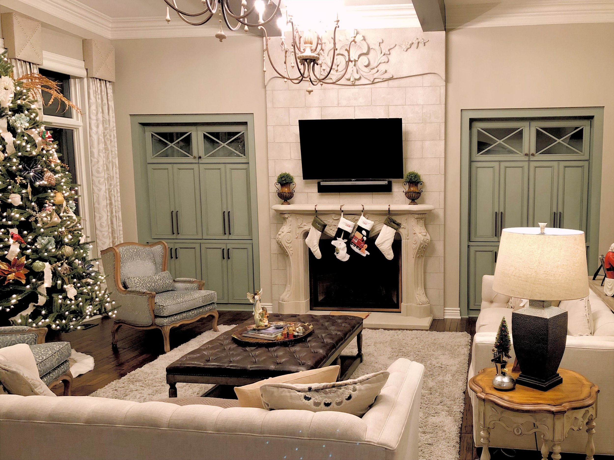 A cozy living room decorated for Christmas with a Christmas tree, stockings hanging from the fireplace, and holiday decorations, featuring a sofa, chairs, a coffee table, and a TV mounted above the fireplace.