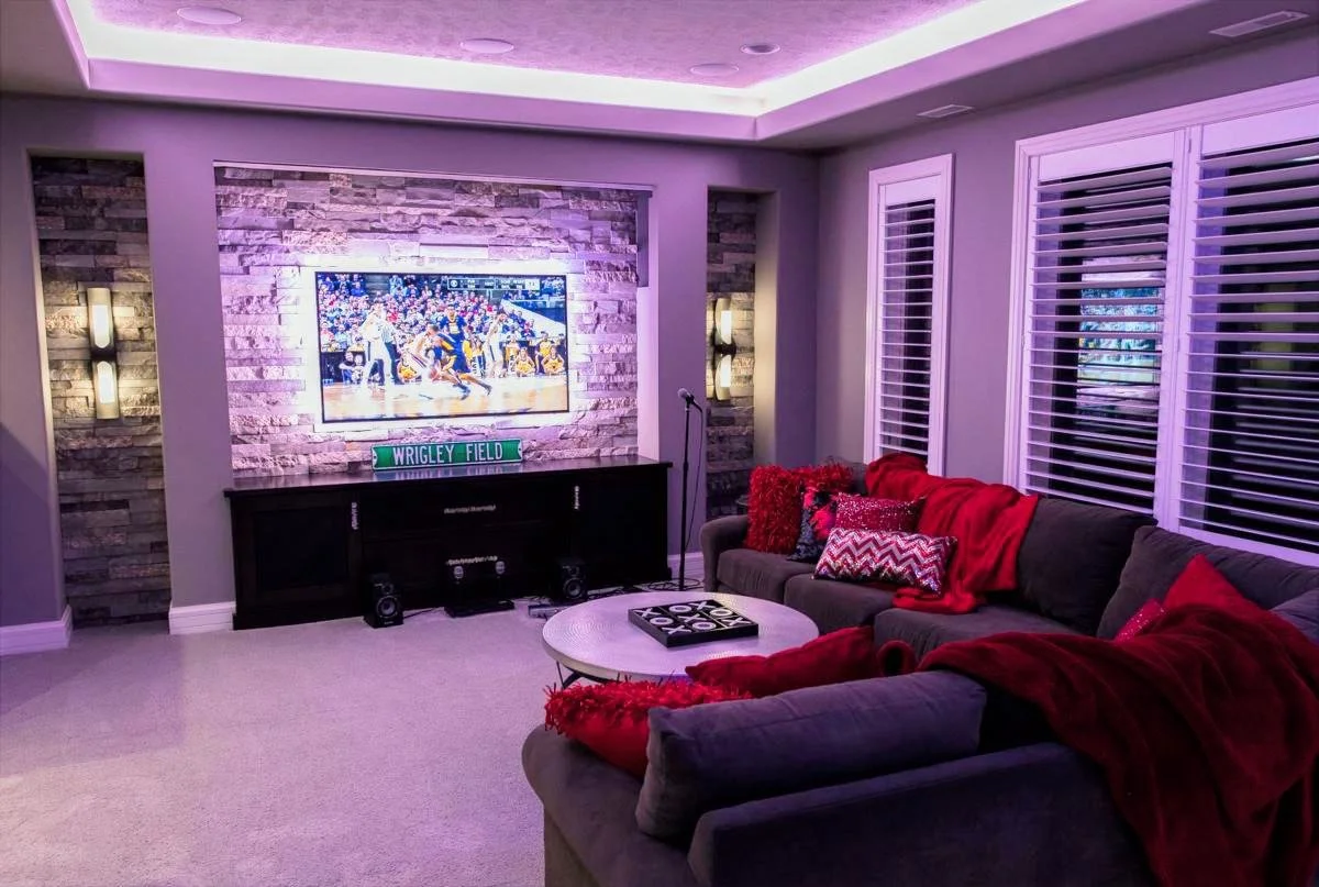 Living room with a large gray sectional sofa with red and patterned pillows and throws, a circular coffee table with a game, a TV mounted on a brick accent wall, and multiple windows with white blinds, illuminated with purple lighting.