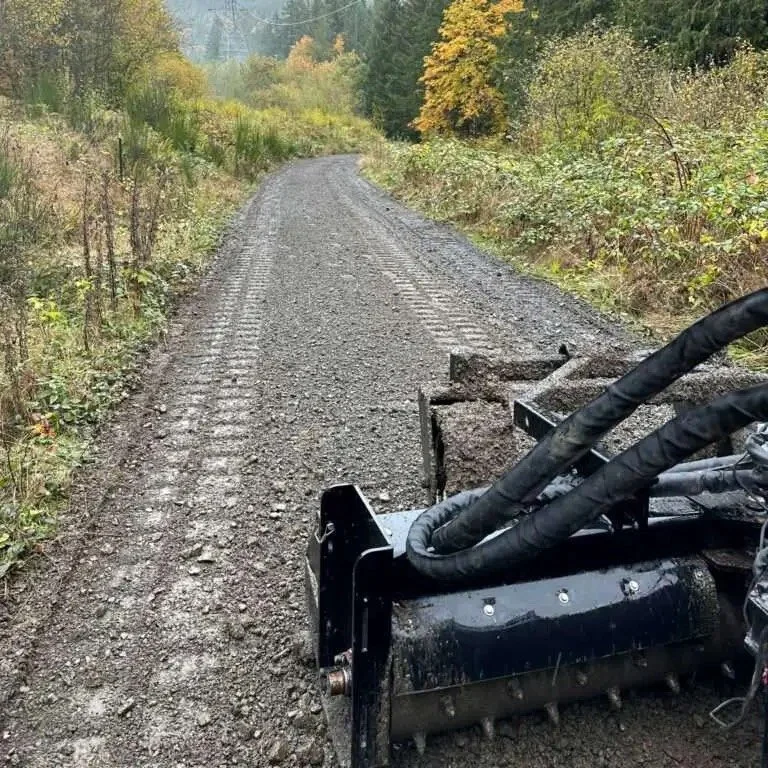 Close-up photo of a piece of heavy machinery working on a gravel road in a forested area, with trees and foliage showing fall colors in the background.
