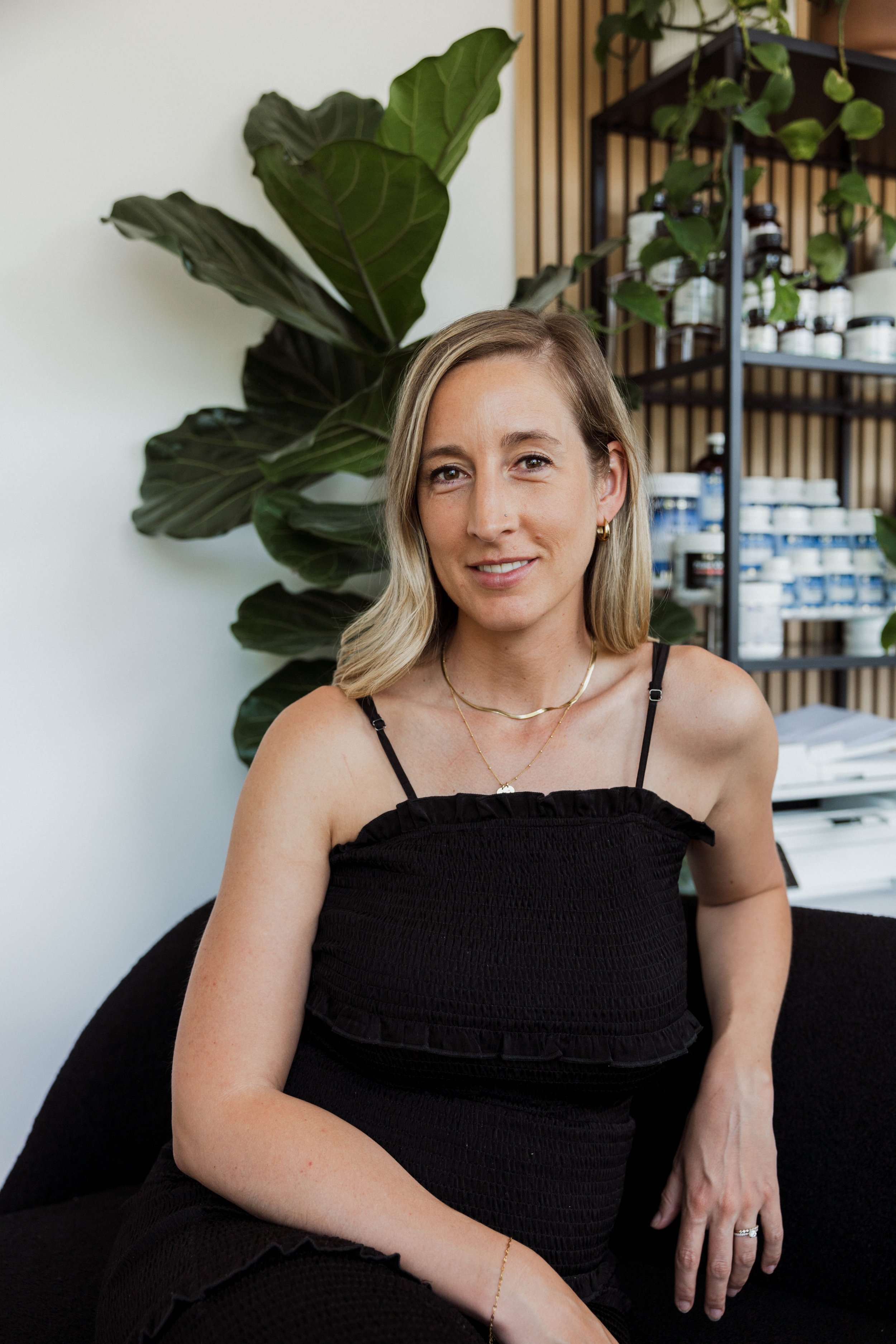 A woman with light skin and light brown hair seated on a black sofa, smiling at the camera, with large green leaves behind her and shelves with supplement bottles and jars in the background.