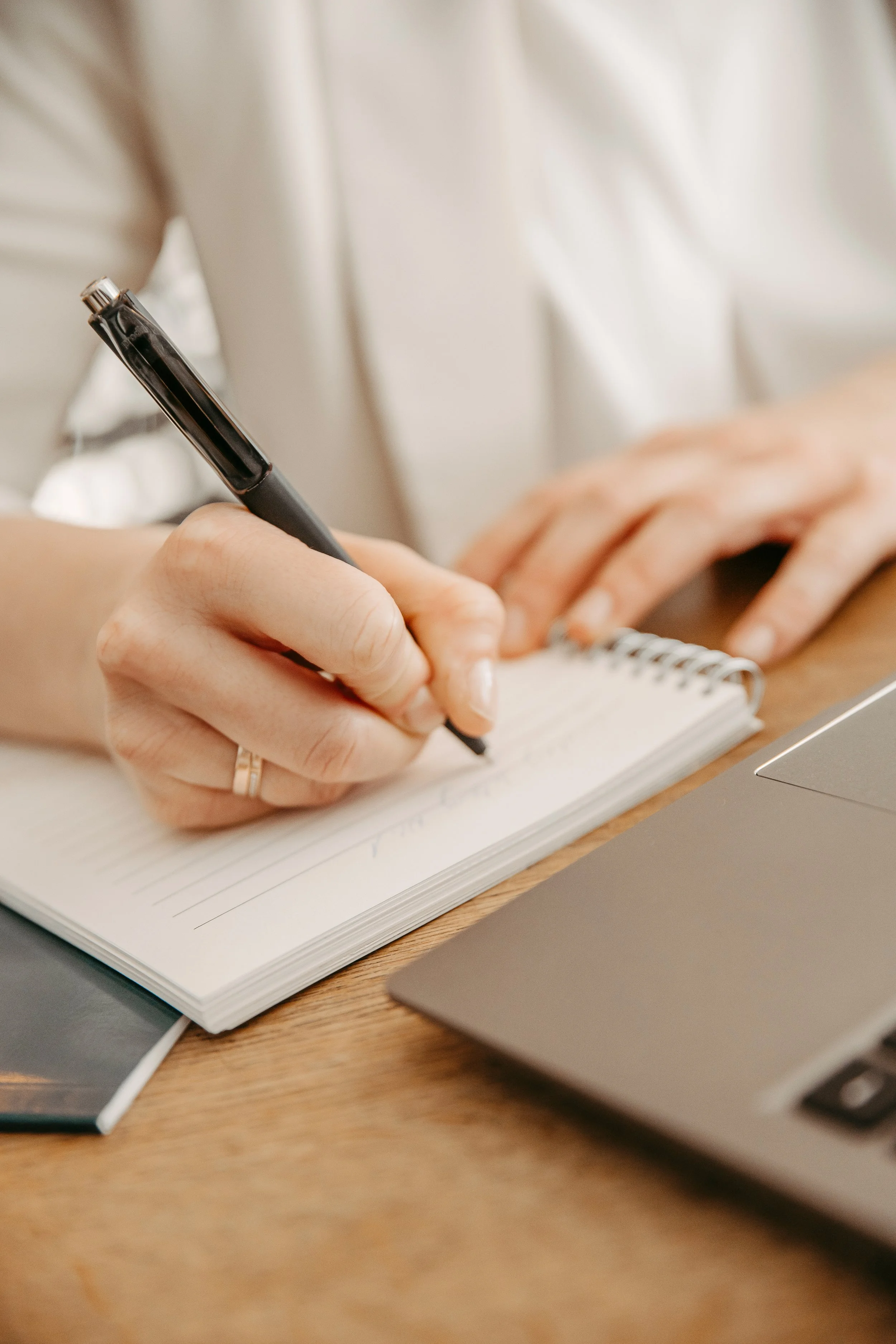 Person schreibt in ein Notizbuch, mit einem Laptop und einem Smartphone auf einem Holztisch.