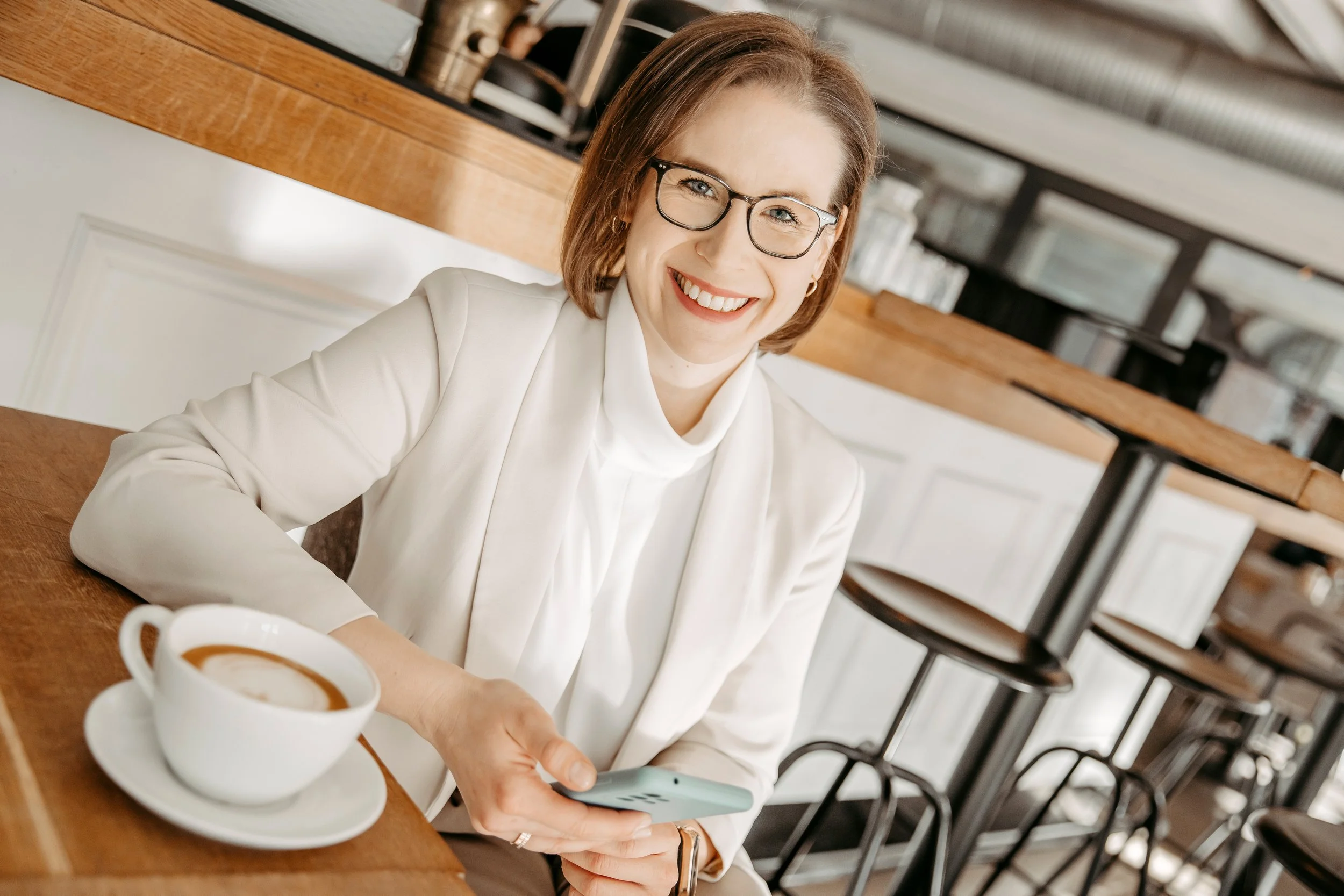 Eine lächelnde Frau mit Brille sitzt in einem Café, trinkt Kaffee und schaut auf ihr Smartphone.