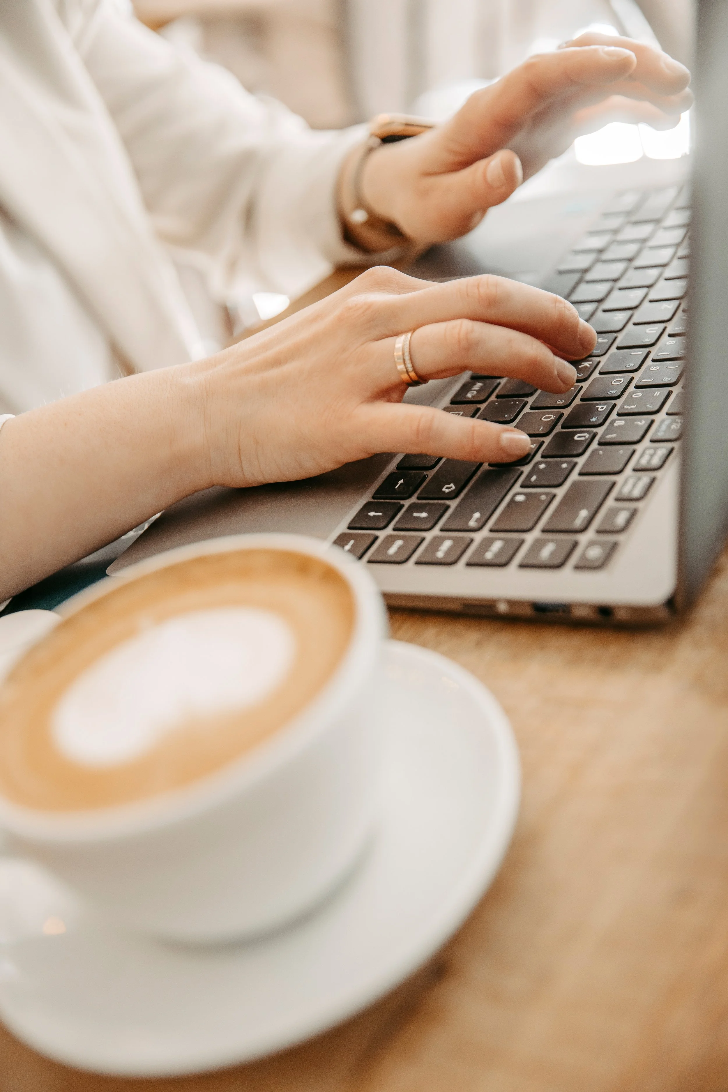 Person tippt auf einer Tastatur, im Vordergrund eine Tasse Kaffee auf einem Unterteller.